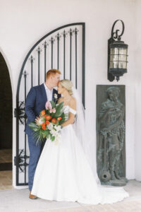 Romantic Bride Holding Tropical Monstera and Palm Frond Leaves, King Protea, Orange Roses, White Orchids, Pincushion Protea, Pink Anthurium Floral Bouquet and Groom Kissing Under Door Arch at Waterfront St. Pete Wedding Venue Hotel Zamora | Tampa Bay Wedding Hair and Makeup Savannah Olivia Beauty