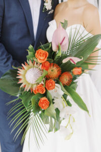 Tropical Pink and Green Wedding | Bride Holding Lush Floral Bouquet with Monstera and Palm Frond Leaves, Orange Roses, Pincushion Protea, King Protea, Pink Anthurium, White Hanging Orchid | Lemon Drops Floral