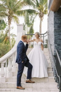 Florida Classic Tropical Elegant Bride Wearing Off the Shoulder A Line Wedding Dress with Groom Kissing Bride Hand On Outdoor Staircase of Waterfront St. Pete Wedding Venue Hotel Zamora | Tampa Bay Wedding Hair and Makeup Savannah Olivia Beauty