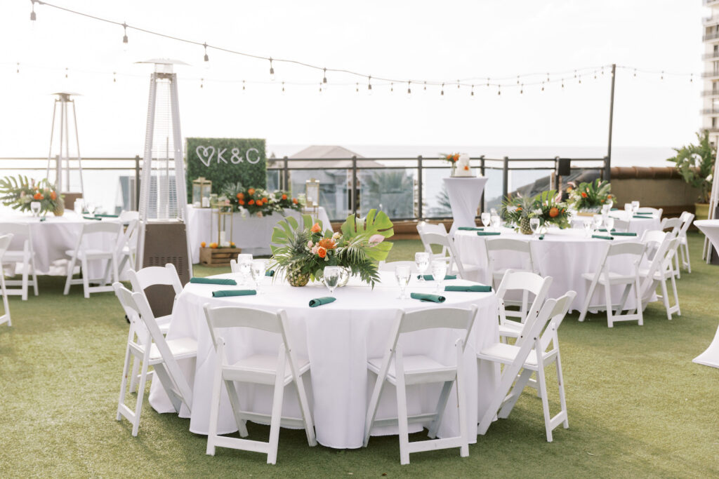 Tropical Pink and Green Rooftop Waterfront Outdoor Wedding Reception Decor, Round Tables with White Linens and Folding Garden Chairs, Teal Napkin, Low Floral Centerpiece, Monstera and Palm Frond Leaves, Orange Roses, Pineapple, String Lights | Tampa Bay Wedding Planner Coastal Coordinating | St. Pete Waterfront Rooftop Wedding Venue Hotel Zamora