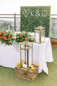 Tropical Pink and Green Wedding Reception Decor | Sweetheart Table with Monstera and Palm Fronds, Orange Roses, White Orchids Floral Arrangement, Gold Lanterns, Wooden Pallet with Lemons, Limes, and Orange Fruits, Greenery Hedge Wall Backdrop with Neon Custom Letters | Tampa Bay Wedding Planner Coastal Coordinating | Waterfront Rooftop Outdoor St. Pete Wedding Venue Hotel Zamora