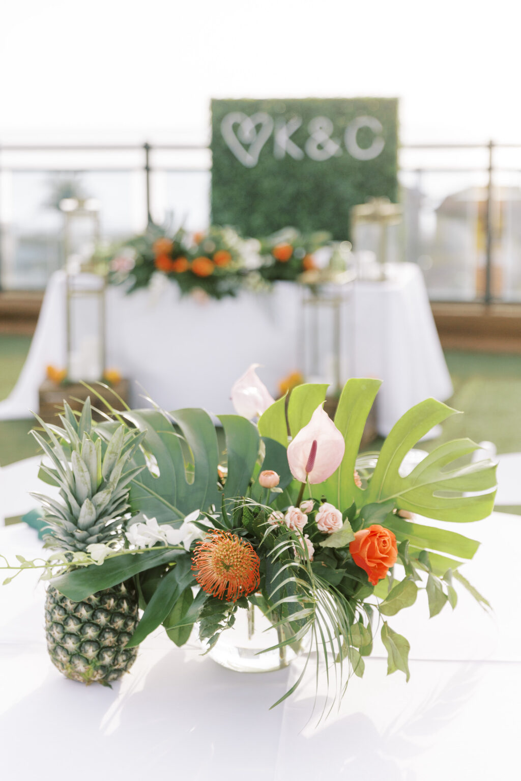 Tropical Pink and Green Rooftop Waterfront Outdoor Wedding Reception Decor, Round Tables with White Linens and Folding Garden Chairs, Teal Napkin, Low Floral Centerpiece, Monstera and Palm Frond Leaves, Orange Roses, Pincushion Protea, Pink Anthurium, Pineapple, String Lights | Tampa Bay Wedding Planner Coastal Coordinating | St. Pete Waterfront Rooftop Wedding Venue Hotel Zamora | Lemon Drops Floral