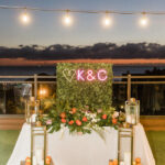 Tropical Pink and Green Wedding Rooftop Outdoor Reception Decor, Greenery Hedge Wall Backdrop with Pink Neon Letters, Sweetheart Table with Monstera and Palm Fronds, Orange Roses, White Orchids Floral Arrangement, Gold Lanterns, Wooden Pallet with Oranges, Limes, and Lemon Fruit, String Lights | St. Pete Rooftop Wedding Venue Hotel Zamora | Tampa Bay Wedding Planner Coastal Coordinating