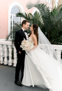 Classic Timeless Bride Wearing A-Line Wedding Dress and Full Length Veil Holding Neutral Floral Bouquet with Groom in Black Tuxedo Outside St. Pete Wedding Venue Historic Pink Palace The Don CeSar Hotel