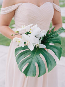 White and Green Modern Minimalist Tropical Wedding, Bridesmaid Holding Champagne Off the Shoulder Dress Holding Monstera Palm Leaf and White Orchid Floral Bouquet