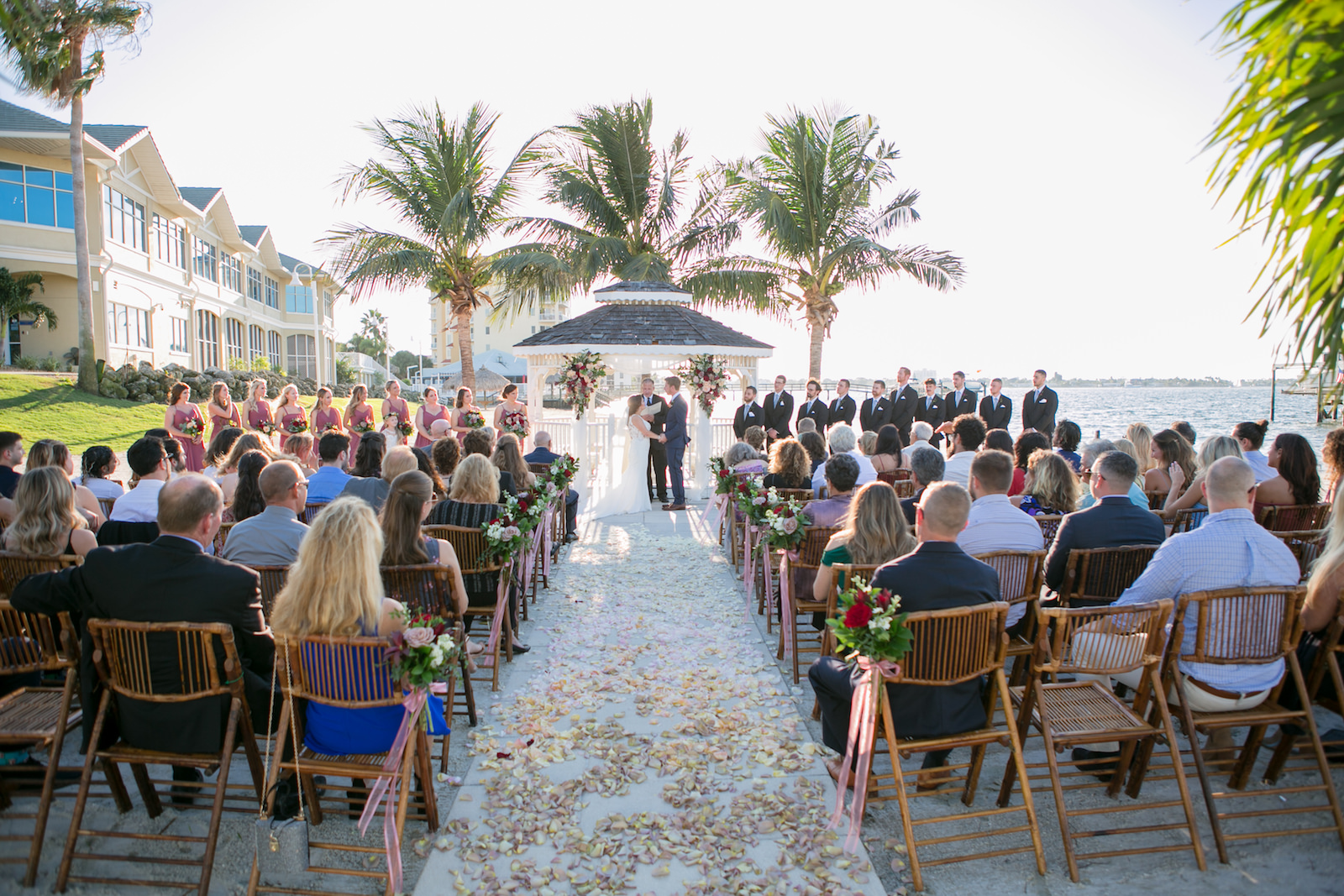 Dusty Rose and Mauve Waterfront Wedding Ceremony Decor, Bride and Groom Exchanging Wedding Vows, Wooden Bamboo Chairs, Tropical Floral Arrangements, Gazebo | Tampa Bay Wedding Photographer Carrie Wildes Photography | St. Pete Wedding Venue Isla Del Sol Yacht and Country Club | Wedding Chair Rentals A Chair Affair | Wedding Planner Perfecting the Plan