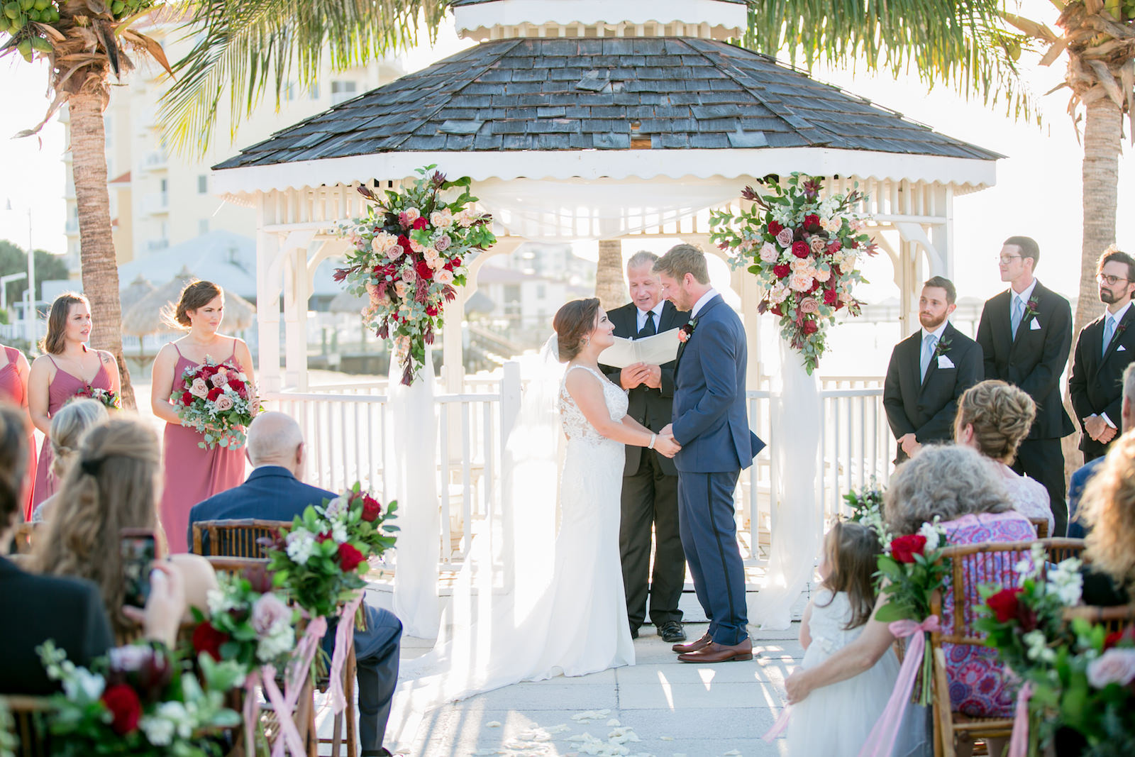 Dusty Rose and Mauve Waterfront Wedding Ceremony Decor, Bride and Groom Exchanging Wedding Vows, Wooden Bamboo Chairs, Tropical Floral Arrangements, Gazebo | Tampa Bay Wedding Photographer Carrie Wildes Photography | St. Pete Wedding Venue Isla Del Sol Yacht and Country Club | Wedding Chair Rentals A Chair Affair | Wedding Planner Perfecting the Plan