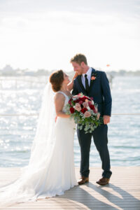 Bride Holding Lush Mauve, Red and White Roses, with Greenery and Eucalyptus Floral Bouquet and Groom Walking on Boat Dock Wedding Portrait | Tampa Bay Wedding Photographer Carrie Wildes Photography | Wedding Florist Iza’s Flowers | St. Pete Wedding Venue Isla Del Sol Yacht and Country Club