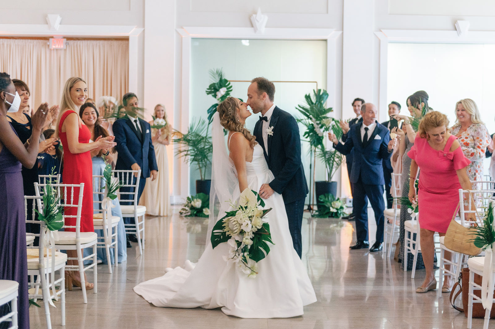 White and Green Modern Minimalist Tropicl Wedding Ceremony, Bride and Groom Kissing After Exchanging Vows Holding Lush Monstera Palm Leaves, White King Protea, Orchids, Anthurium Floral Bouquet | Tampa Bay Wedding Planner Taylored Affairs | Downtown Tampa Historic Wedding Venue The Vault