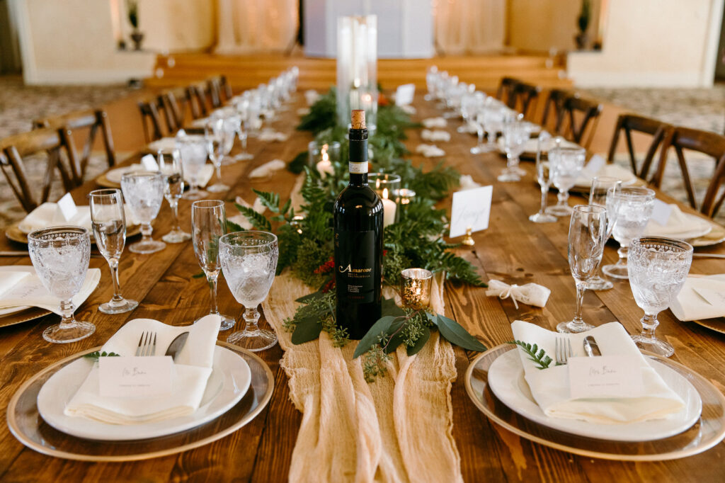 Rustic Italian Elegant Wedding Reception Decor, Wooden Cross Back Chairs, Ivory Cheesecloth Table Runner, Greenery, Candles, Bottle of Wine, Gold Rimmed Chargers, Long Wooden Feasting Table | Tampa Bay Wedding Rentals Outside the Box Rentals