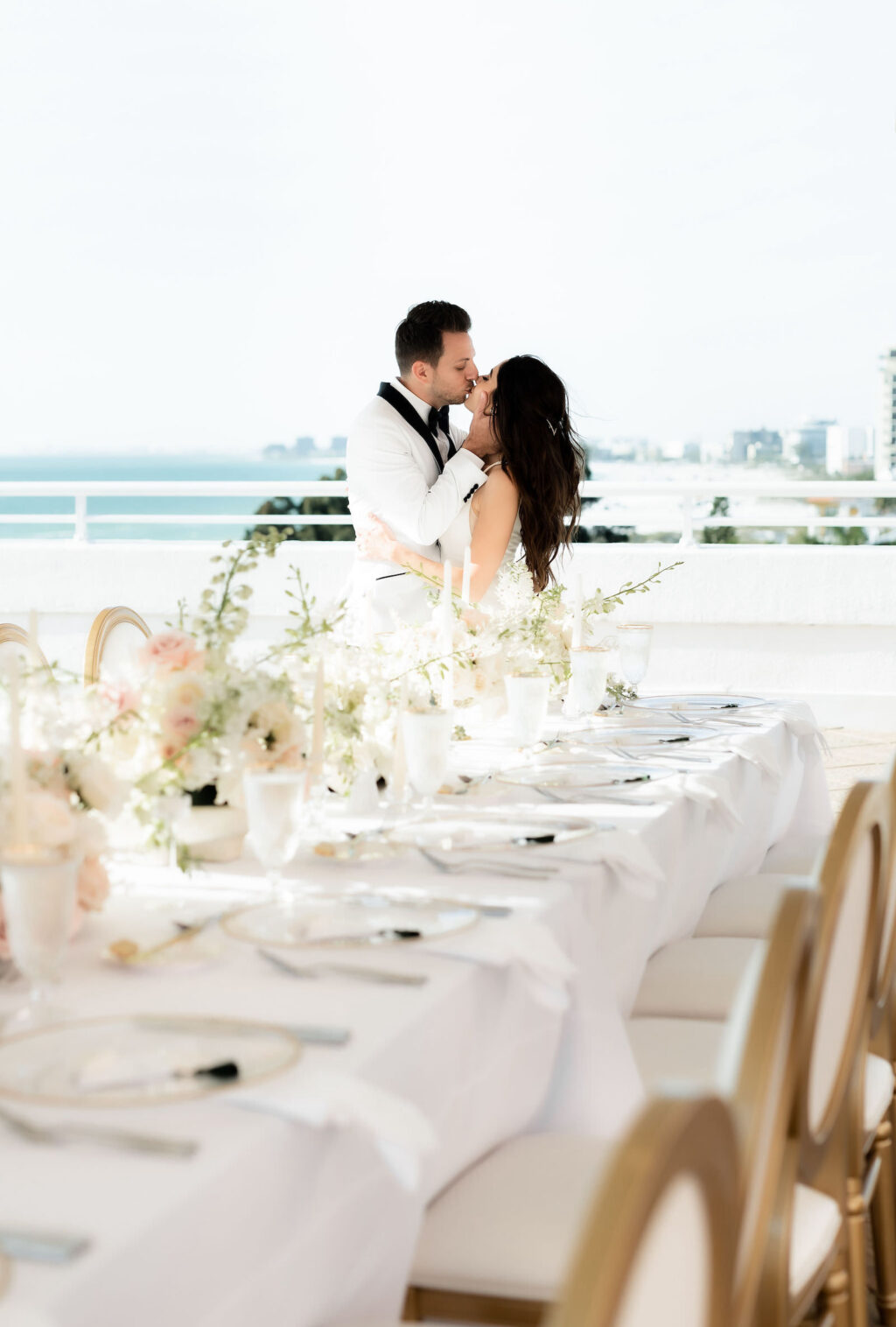 Classic Elegant Bride and Groom in White Tuxedo with Black Bowtie Standing at Wedding Reception Table with Elegant Ivory and Gold Dining Chairs, Low Floral Centerpieces, Rooftop Waterfront St.Pete Historic Wedding Venue The Don CeSar Hotel | Tampa Bay Wedding Planner Elegant Affairs by Design | Wedding Rentals Kate Ryan Event Rentals