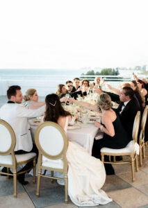 Classic Timeless Bride and Groom Sitting at Elegant Gold and White Dining Chairs Cheering with Guests during Small Intimate Wedding Reception at Long Table | Tampa Bay Wedding Planner Elegant Affairs by Design | Wedding Rentals Kate Ryan Event Rentals | Historic St. Pete Rooftop Waterfront Wedding Venue The Don CeSar Hotel