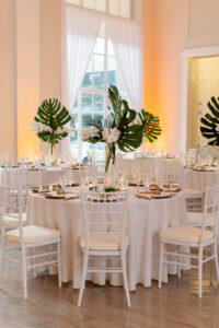 White and Green Modern Minimalist Tropical Wedding Reception Decor, White Chiavari Chairs, Gold Chargers and Silverware, Tall Clear Hurricane Vases with Monstera Palm Leaves and White Orchid Centerpiece | Tampa Bay Wedding Planner Taylored Affairs | Downtown Tampa Historic Wedding Venue The Vault