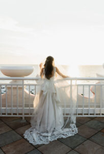 Romantic Timeless Sunset Bride Overlooking Balcony of Waterfront Rooftop Historic St. Pete Wedding Venue The Don CeSar Hotel Wearing Classic Wedding Dress with Bow in the Back and Full Length Lace Veil