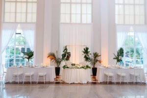 White and Green Modern Minimalist Tropical Wedding Reception Decor, White Chiavari Chairs, Gold Chargers and Silverware, Tall Clear Hurricane Vases with Monstera Palm Leaves and White Orchid Centerpiece, White Linen Backdrop Sweetheart Table, Bridal Party Tables, Planted Palm Fronds, Gold Arch | Tampa Bay Wedding Planner Taylored Affairs | Downtown Tampa Historic Wedding Venue The Vault