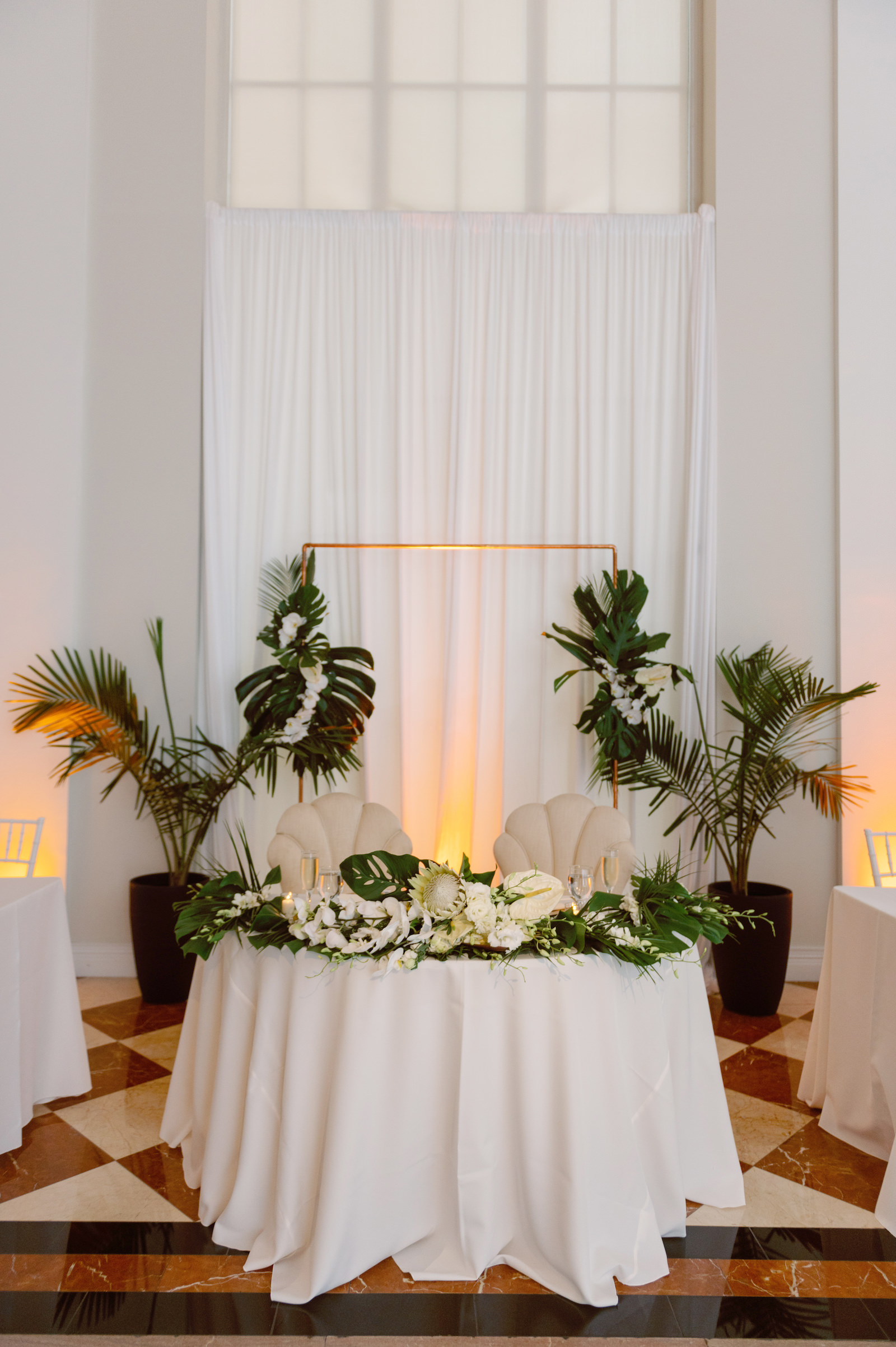 White and Green Modern Minimalist Tropical Wedding Reception Decor, Sweetheart Table with Seashell Tufted Dining Chairs, Palm Fronds, Monstera Leaves, White King Protea, Roses, Orchids, Anthurium Floral Arrangements, Planted Palm Fronds in Pots, Gold Arch, Linen Draping Backdrop | Tampa Bay Wedding Planner Taylored Affairs | Downtown Tampa Wedding Venue The Vault
