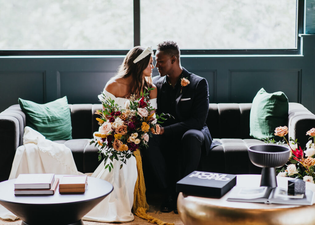Modern and Edgy Fall Wedding, Tampa Bride Wearing Off the Shoulder Wedding Dress Holding Yellow, Burgundy Red and Blush Pink Roses Floral Bouquet with Greenery, Groom Wearing All Black Tuxedo and Peach Rose Boutonniere Sitting on Velvet Couch | Tampa Bay Wedding Venue Hyde House