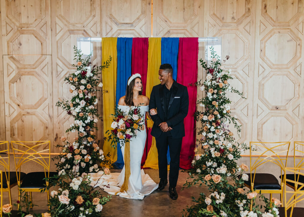Modern and Edgy Fall Wedding Decor, Bride and Groom Exchanging Wedding Ceremony Vows, Chic Gold Chairs, Greenery and Blush Pink, White and Mauve Roses Floral Arrangements Two Part Standing Floral Ceremony Arch, Primary Colors Yellow, Blue and Red Linen Draping | South Tampa Wedding Venue Hyde House | Wedding Rentals Kate Ryan Event Rentals