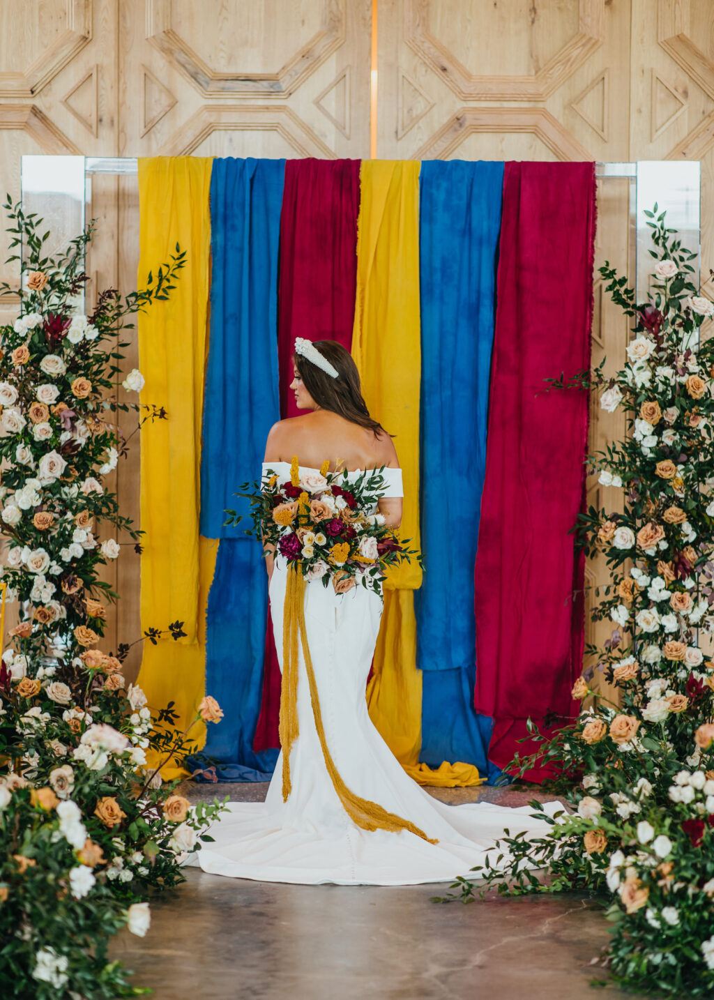 Modern and Edgy Fall Wedding Decor, Bride Wearing Off the Shoulder Fitted Wedding Dress Holding Burgundy Red, and Yellow Floral Bouquet with Mustard Yellow Linen Hanging, Chic Gold Chairs, Greenery and Blush Pink, White and Mauve Roses Floral Arrangements Two Part Standing Floral Ceremony Arch, Primary Colors Yellow, Blue and Red Linen Draping | South Tampa Wedding Venue Hyde House
