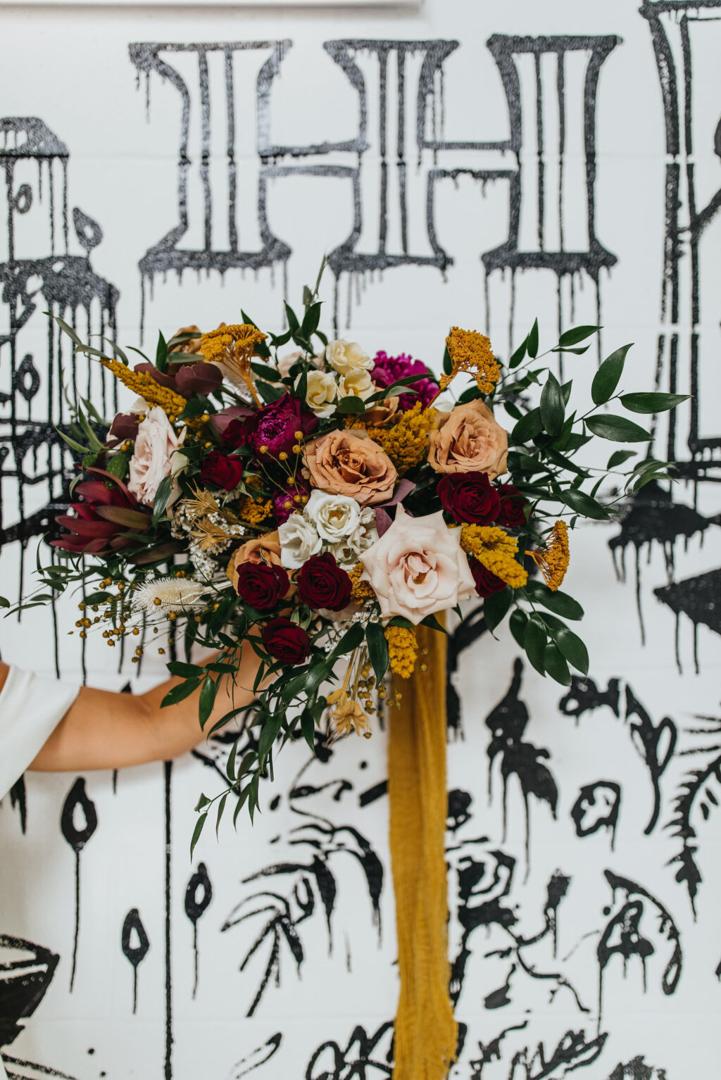 Modern and Edgy Fall Wedding, Bride Holding Wild Blush Pink, Burgundy Red Roses, Yellow Flowers and Greenery Bouquet with Mustard Yellow Hanging Linen, Black and White Graffiti Wall | South Tampa Wedding Venue Hyde House