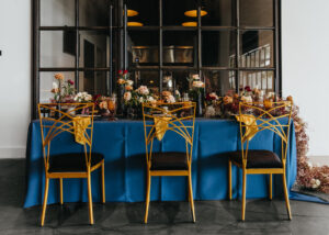 Modern and Edgy Fall Wedding Decor, Long Table with Royal Blue Table Linen, Lush Floral Table Runner, Chic Gold Chairs | Tampa Bay Wedding Venue Hyde House | Wedding Rentals Kate Ryan Event Rentals
