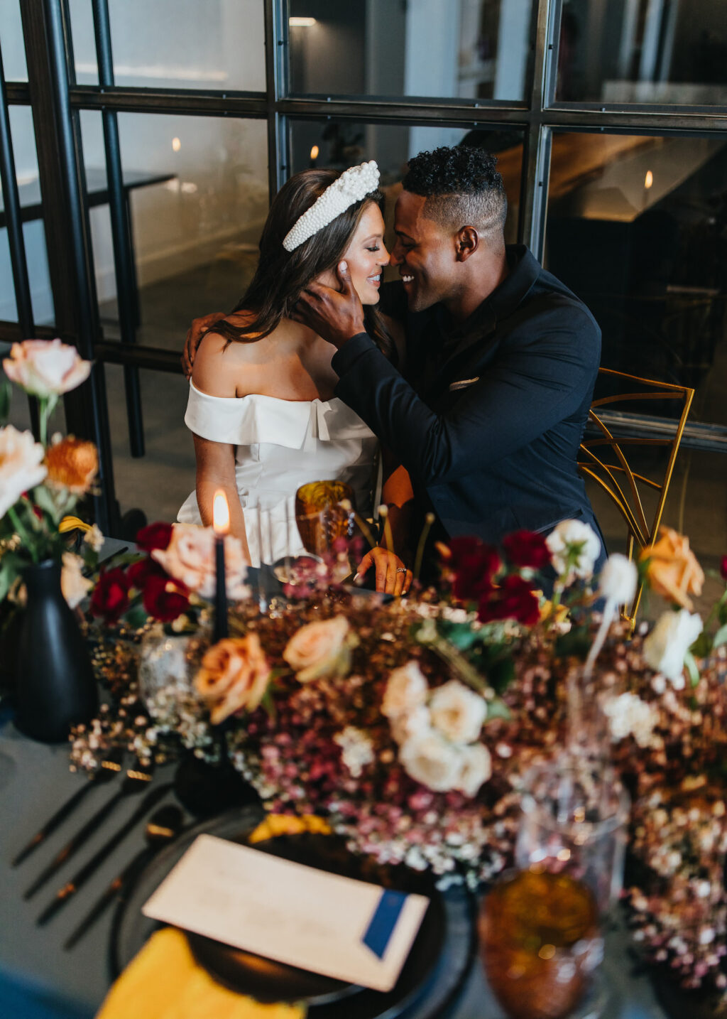 Modern and Edgy Fall Wedding Reception, Groom Wearing All Black Tuxedo Intimate Sitting at Reception Table with Bride Wearing Off the Shoulder Wedding Dress, Thick Pearl Headband,Burgundy Red, White, Blush Pink and Yellow Roses with Greenery Flowers Babys Breathe Floral Centerpiece | Tampa Bay Wedding Venue Hyde House