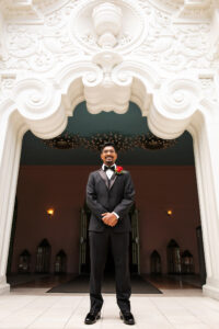 Groom Wearing Black Tuxedo, Bow Tie and Red Rose Boutonierre Standing Under Archway of St. Petersburg Wedding Venue The Vinoy Renaissance