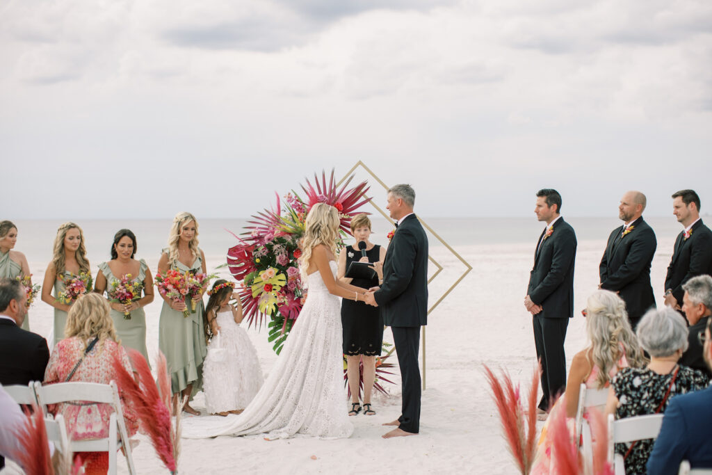 Vibrant Boho Beach Wedding, Bride and Groom Exchanging Wedding Vows, Gold Geometric Diamond Arch with Vibrant Hot Pink and Green Monstera Leaves, Palm Fronds and Roses Floral Arrangement | Tampa Bay Wedding Officiant A Wedding with Grace