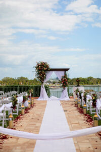 Elegant Garden Waterfront Wedding Ceremony Decor, Large Floral Vases with Red, Pink and White Roses with Greenery Floral Arrangements, White Drapery, White Folding Garden Chairs, Red Rose Petals, White Linen Aisle Runner, Wooden Arch with Floral Arrangements and White Draping | Tampa Bay Wedding Photographer Limelight Photography | Wedding Planner Breezin Weddings | Wedding Florist Lemon Drops | Wedding Rentals Outside the Box Event Rentals