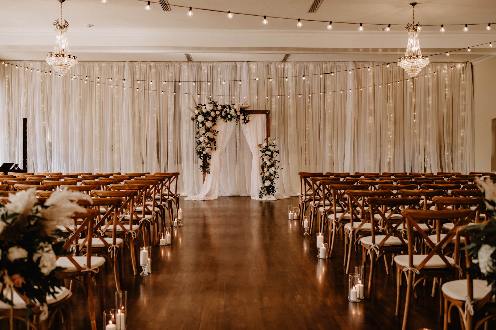 Neutral Boho Modern Wedding Ceremony Decor, Wooden Cross Back Chairs, Hanging String Lights, Wooden Arch with White Linens, Lush Greenery, White Flowers and Pampas Grass Floral Arrangements | Tampa Bay Wedding Planner Taylored Affairs | Wedding Rentals Gabro Event Services