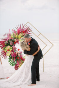 Vibrant Boho St. Pete Beach Wedding Ceremony Decor, Groom Dip Kissing Bride, Gold Geometric Diamond Arch with Hot Pink and Vibrant Green Monstera Leaves, Palm Fronds and Roses Floral Arrangement