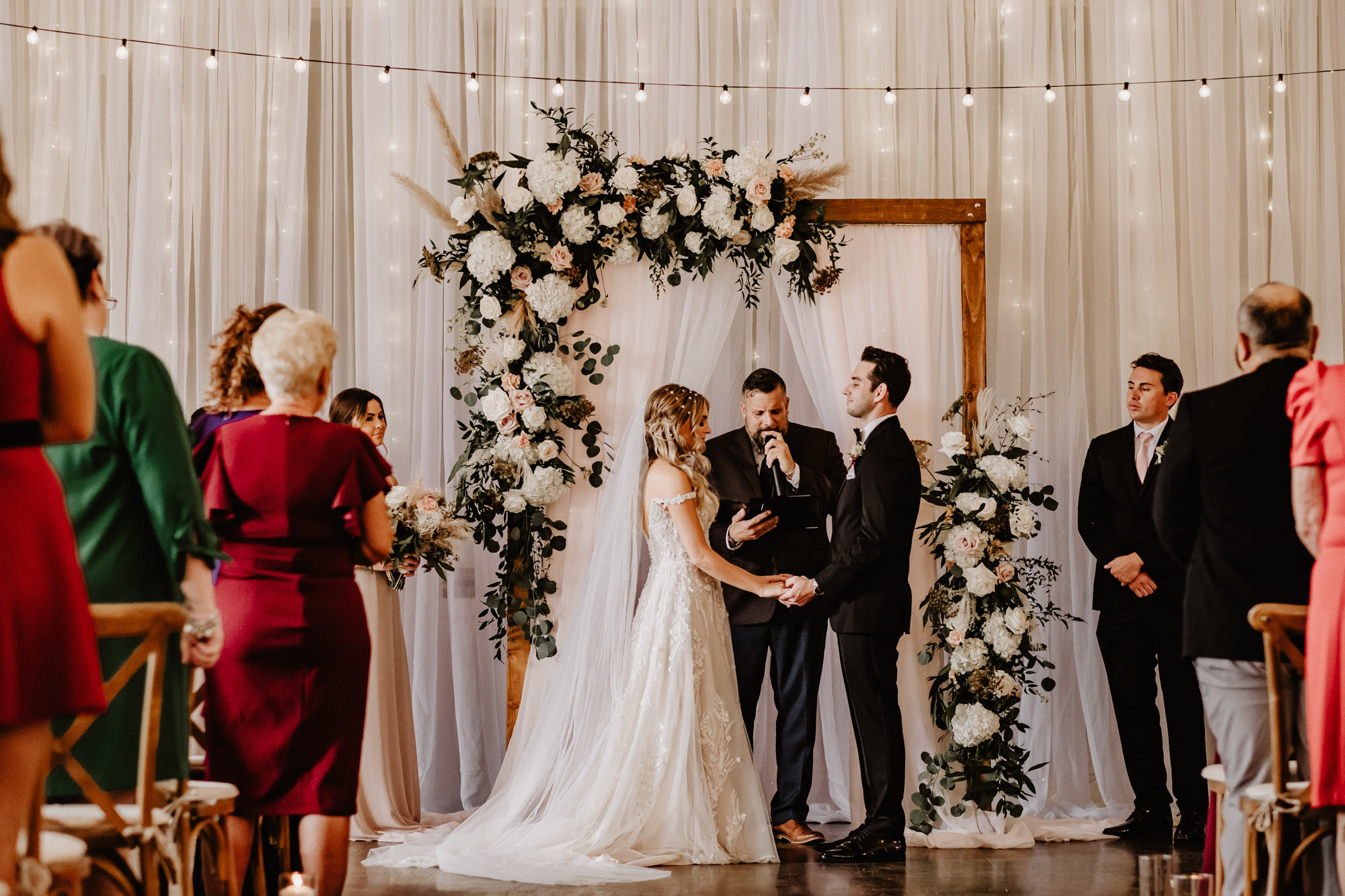 Bride and Groom Exchanging Wedding Vows During Neutral Boho Modern Wedding Ceremony Decor, Wooden Cross Back Chairs, Hanging String Lights, Wooden Arch with White Linens, Lush Greenery, White Flowers and Pampas Grass Floral Arrangements | Tampa Bay Wedding Planner Taylored Affairs | Wedding Rentals Gabro Event Services