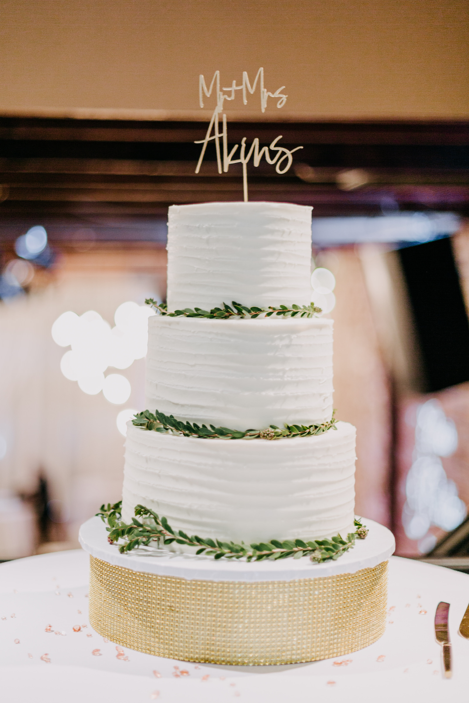 Four Tier White Textured Wedding Cake With Gold Details, Greenery, and a Gold Cake Topper