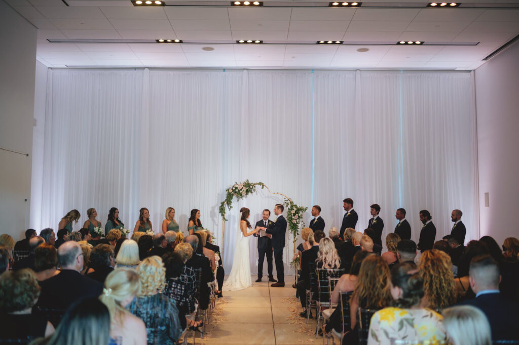 Modern Minimalist Bride and Groom Exchanging Wedding Vows During Ceremony, White Draping, Unique Arch with Lush Greenery and White, Blush Pink Roses Floral Arrangements