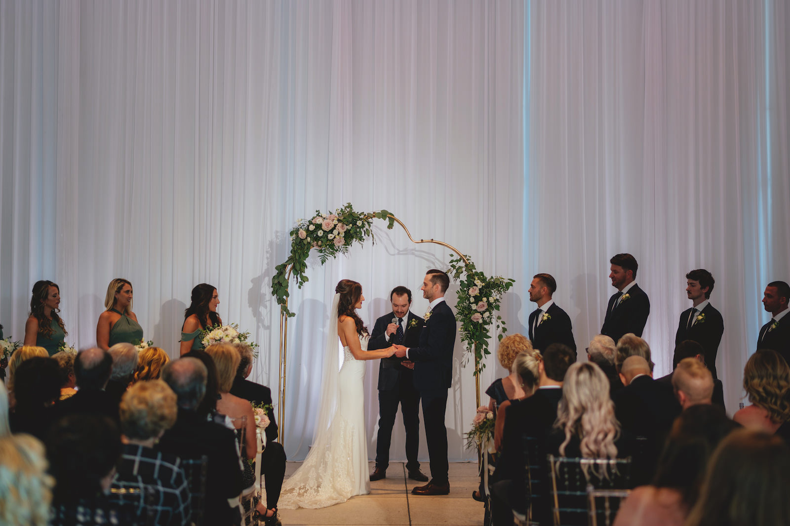 Modern Minimalist Bride and Groom Exchanging Wedding Vows During Ceremony, White Draping, Unique Arch with Lush Greenery and White, Blush Pink Roses Floral Arrangements