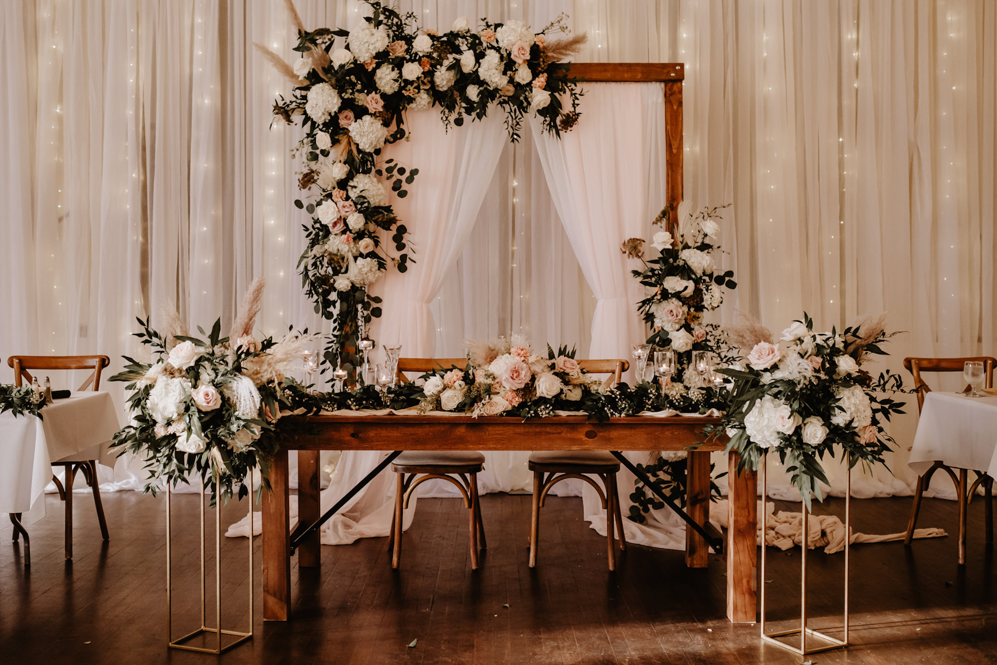 Neutral Boho Modern Wedding Reception Decor, Wooden Sweetheart Table, Wooden Cross Back Chairs, Wooden Arch with White Draping, Hanging String Lights Backrop, Lush Greenery, White Hydrangeas, Blush Pink Roses, Pampas Grass Flowers | Tampa Bay Wedding Planner Taylored Affairs | St. Pete Wedding Rentals Gabro Event Services