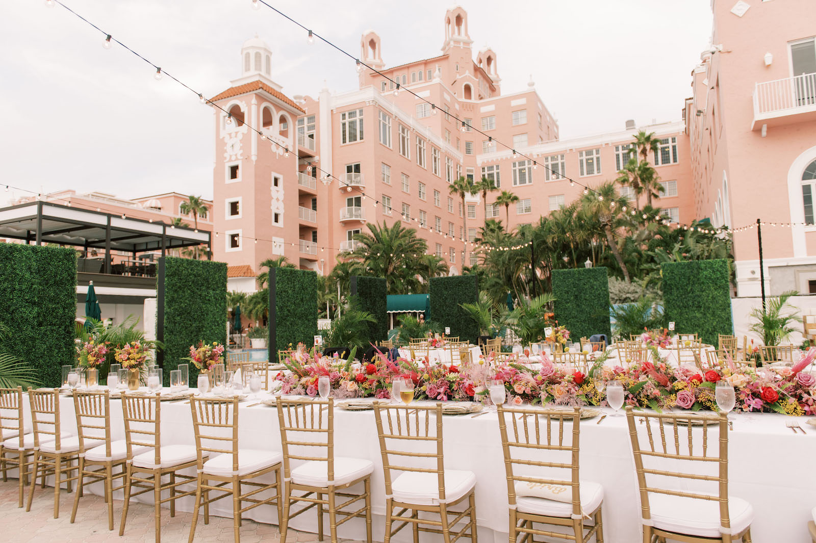 Vibrant Boho Wedding Reception Decor, Wedding Party Head Table, Gold Chiavari Chairs, Low Hot Pink and Red Roses, Pampas Grass Low Floral Centerpieces, Hanging String Lights, Greenery Hedge Wall | St. Petersburg Historic Wedding Venue The Don CeSar