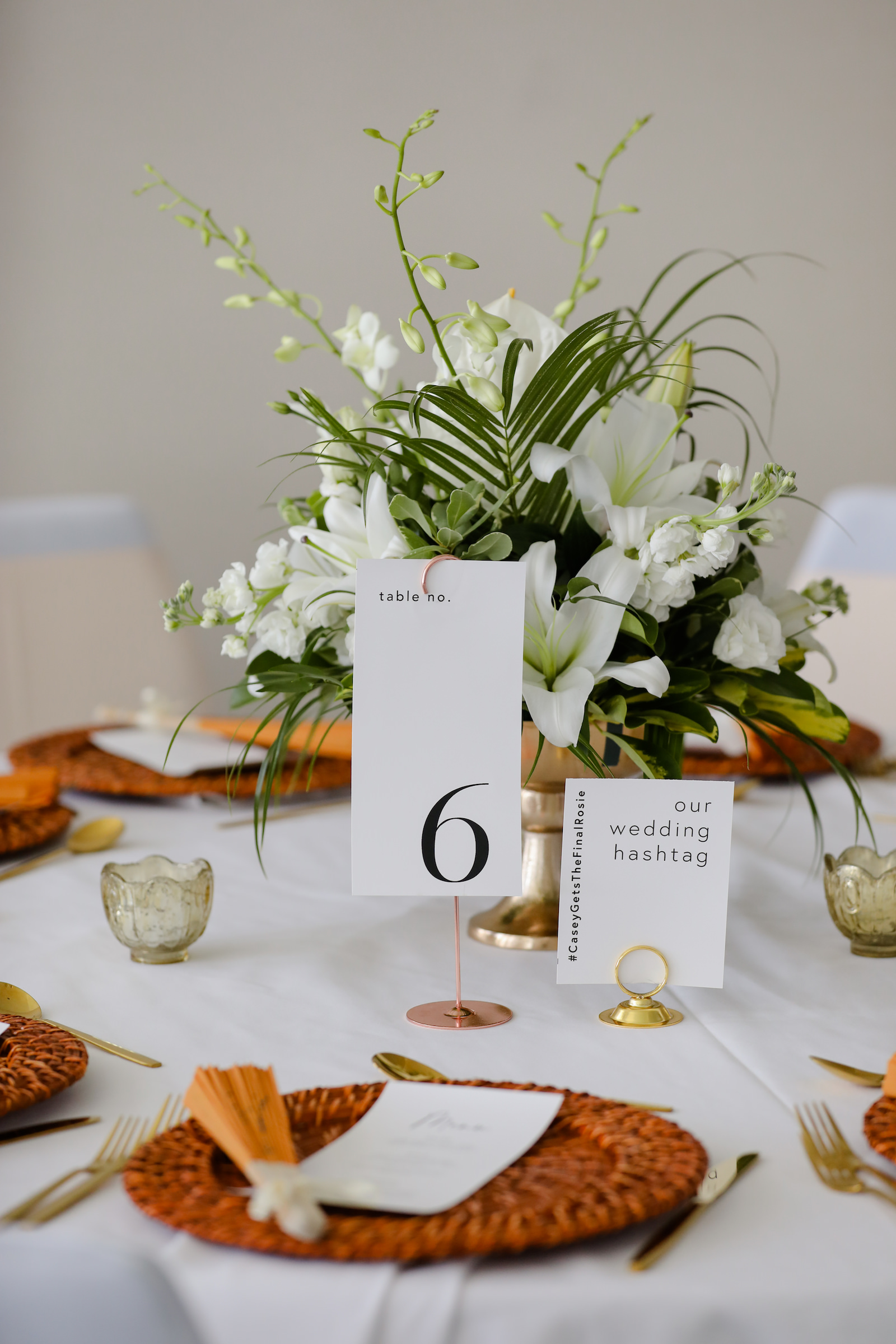 Modern Tablescape and Table Numbers with Tall Gold Centerpieces with Greenery and White Flowers and Woven Chargers