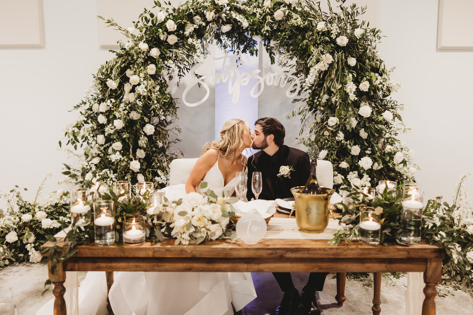 Classic Wedding Reception Decor, Bride and Groom Sitting at Long Wooden Sweetheart Table, Ivory Tufted Loveseat, Lush White Roses and Greenery Floral Arch, Custom Neon Sign, Floating Candles