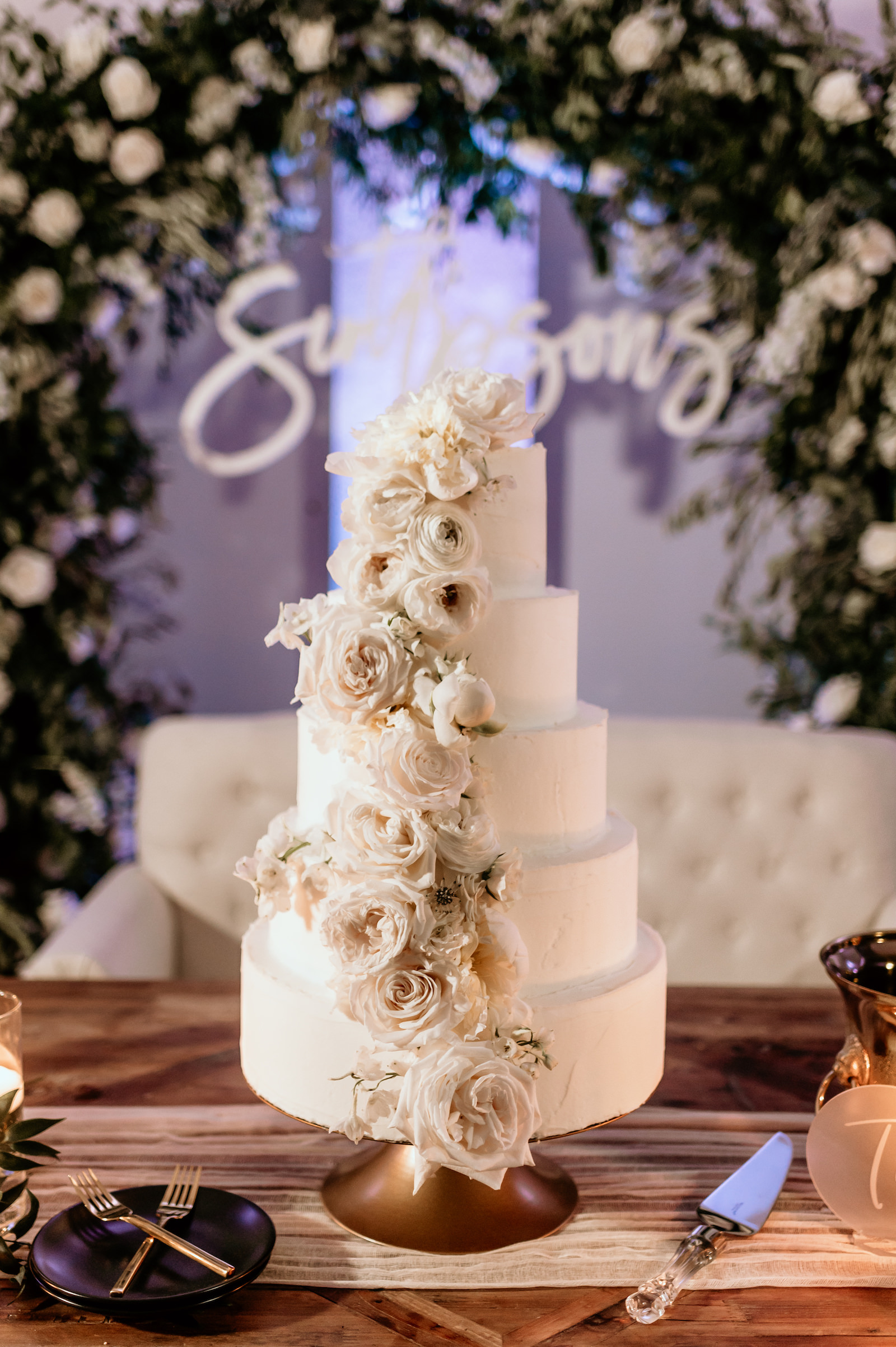 Five Tier White Classic Wedding Cake with Cascading White Flowers on Wooden Sweetheart Table, Lush White Roses and Greenery Floral Arch