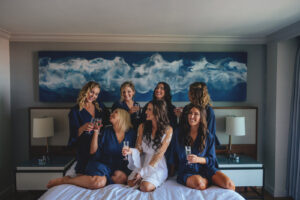 Modern Minimalist Bride Wearing White Robe and Bridesmaids in Navy Blue Robes Drinking Champagne on Bed Getting Wedding Ready