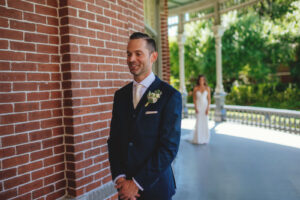Groom Waiting for First Look Wearing Blue Suit and White Tie with White Floral Boutonniere, Bride in the Background