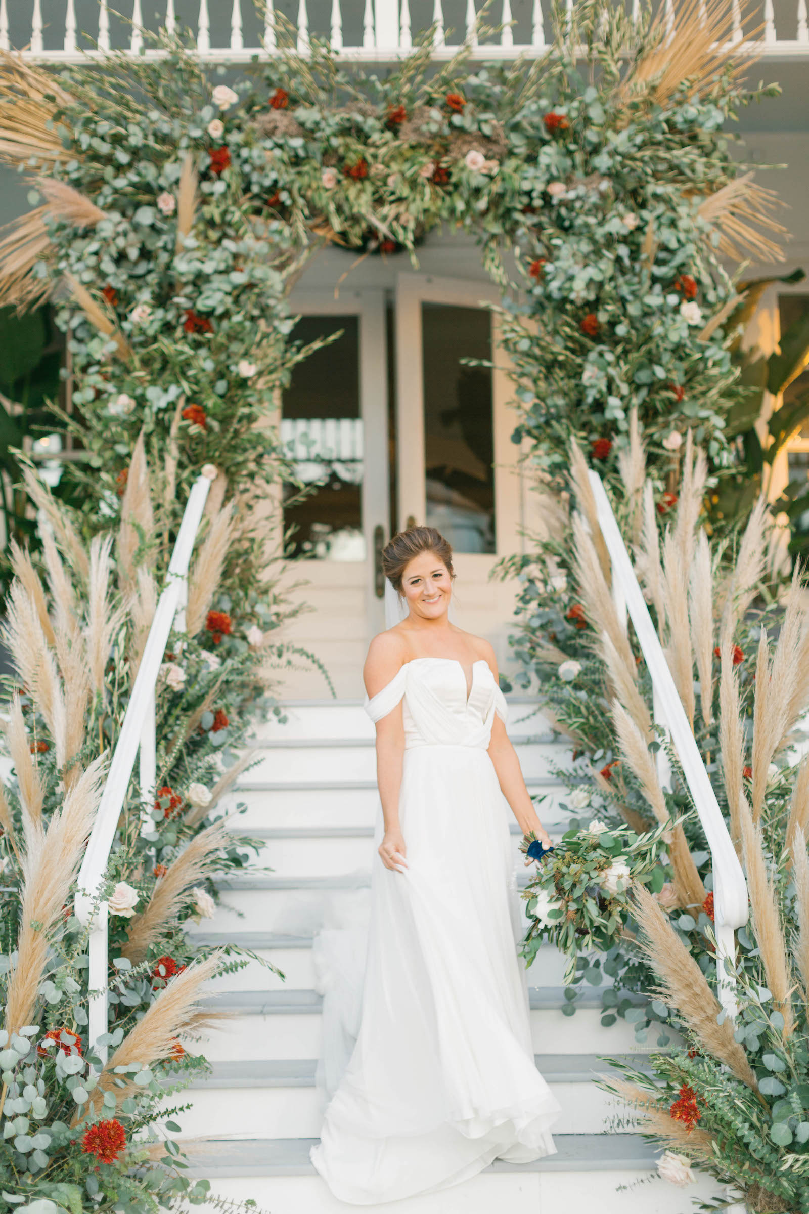 Boho Garden Wedding Ceremony Decor, Lush Greenery Eucalyptus, Dried Palm Leaves and Red, Blush Pink Floral Arch with Pampas Grass Arrangements, Bride Standing On Staircase of Florida Mansion Wedding Venue Belleview Inn | Tampa Bay Wedding Planner Parties A'la Carte