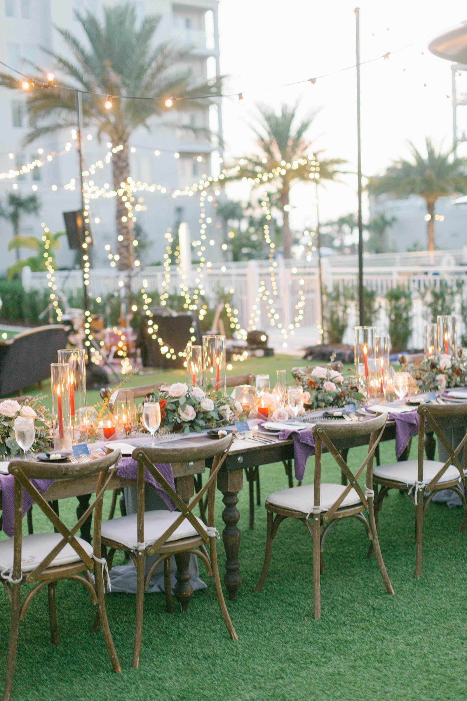 Boho Garden Wedding Reception Decor, Long Wooden Feasting Table with Wooden Cross Back Chairs, String Lights, Purple Linen Table Runner with Greenery Garland, Red Candlesticks, Gold Geometric Vases, White Roses | Tampa Bay Wedding Planner Parties A'la Carte | Florida Wedding Venue Belleview Inn