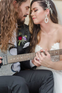 Modern Rock and Roll Wedding, Groom Wearing Custom Black Leather Suit Jacket Playing Black Guitar, Bride Wearing Strapless Ballgown Wedding Dress Sitting on Couch, Round Gold Circular Arch with Jewel Tone Red and Purple Flowers with Greenery Arrangement | Tampa Bay Wedding Photographer Amanda Zabrocki Photography | Wedding Florist Save the Date Florida | Wedding Rentals Kate Ryan Event Rentals