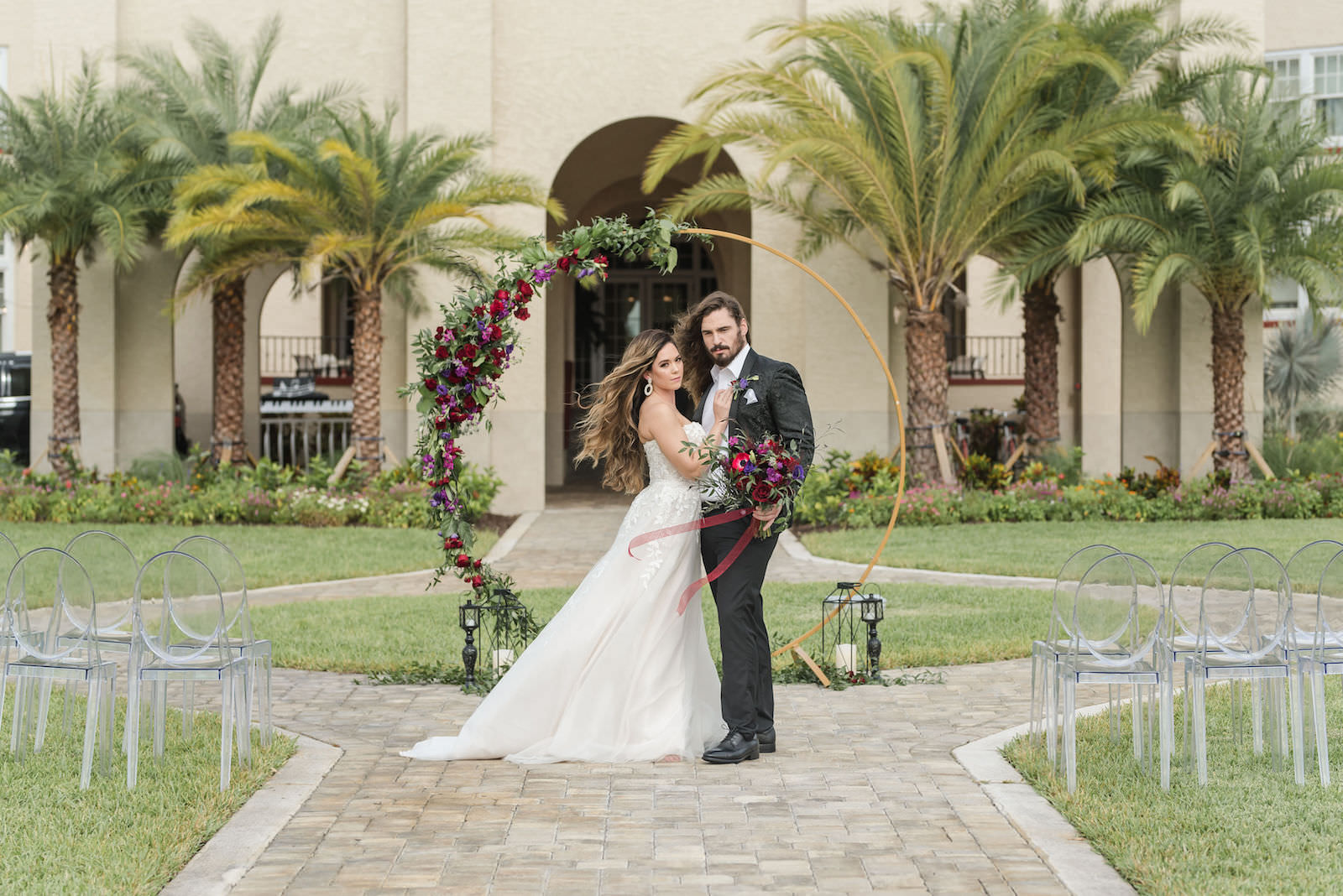 Modern Rock N Roll Wedding, Bride and Groom Standing Outside, Gold Metal Round Arch with Jewel Tone Red and Purple Flowers with Greenery Floral Arrangements, Acrylic Ghost Chairs | Tampa Bay Wedding Photographer Amanda Zabrocki Photography | Wedding Rentals Kate Ryan Event Rentals | Dunedin Wedding Venue The Fenway