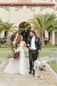 Modern Rock N Roll Wedding, Bride and Groom with Dog Walking Outside, Gold Metal Round Arch with Jewel Tone Red and Purple Flowers with Greenery Floral Arrangements, Acrylic Ghost Chairs | Tampa Bay Wedding Photographer Amanda Zabrocki Photography | Wedding Rentals Kate Ryan Event Rentals | Dunedin Wedding Venue The Fenway | Wedding Pet Planner FairyTail Pet Care