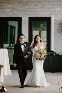 Modern Boho Bride Wearing Fitted Lace Bodice Wedding Dress Holding Blush Pink and Rust Orange Roses with Greenery Floral Bouquet, Walking with Dad During Processional Wedding Ceremony Photo | Tampa Bay Wedding Florist Iza's Flowers | Wedding Hair and Makeup Adore Bridal