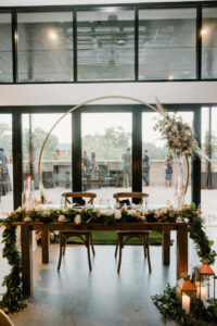 Modern Boho Wedding Reception Decor, Sweetheart Table, Long Wooden Table with Wooden Crossback Chairs, Long Greenery Table Runner with Roses, Round Gold Metal Arch with Lush Floral Arrangement, Lanterns | Tampa Bay Wedding Florist Iza's Flowers | Unique South Tampa Wedding Venue Hyde House