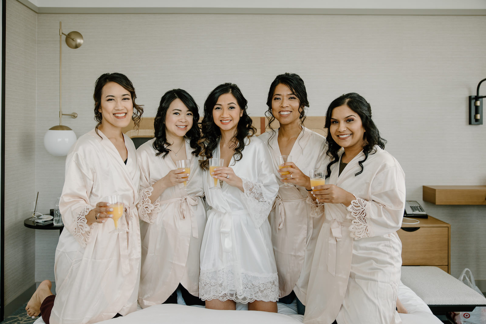 Modern Boho Bride Getting Wedding Ready with Bridesmaids in Matching Ivory Robes Drinking Champagne on Bed | Tampa Bay Wedding Hair and Makeup Adore Bridal
